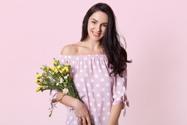 Isolated shot of cheerful dark haired woman wears polka dot summer dress, holds bouquet of flowers, shows bare shoulders