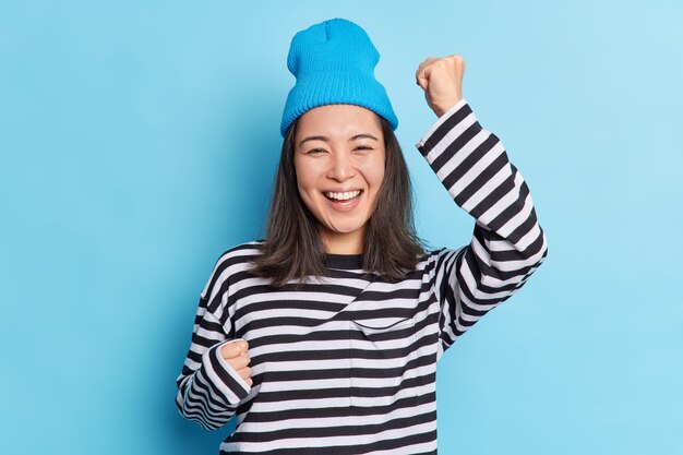 Isolated shot of cheerful happy Asian girl raises hands dances carefree celebrates success smiles broadly expresses sincere emotions and happines