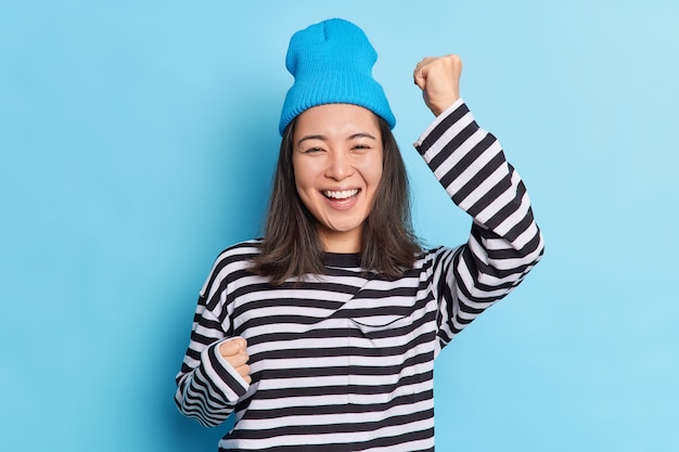 Isolated shot of cheerful happy Asian girl raises hands dances carefree celebrates success smiles broadly expresses sincere emotions and happines