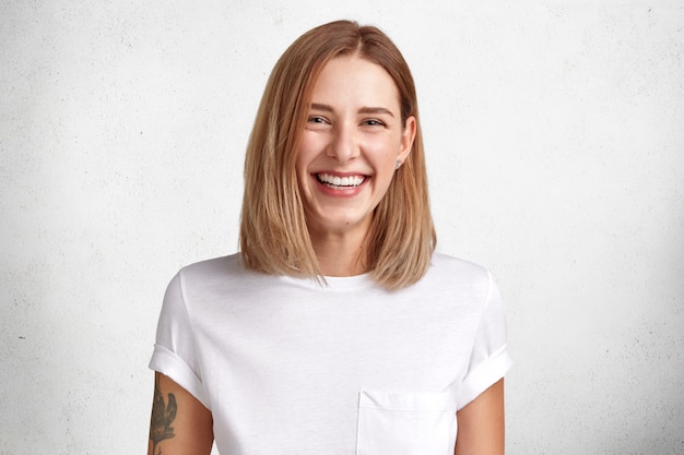 Isolated shot of cheerful satisfied young female with pleasant look, broad chaming smile, tattooed arm, dressed in casual whte t shirt, poses in studio, glad to achieve success at work and life