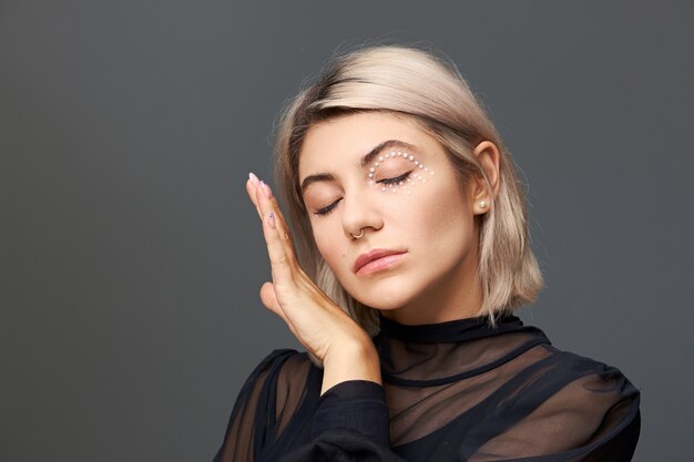 Isolated shot of cute charming young twenty year old woman in transparent black blouse keeping eyes closed and touching gently her soft skin wearing nose ring, stylish haircut and bright make up