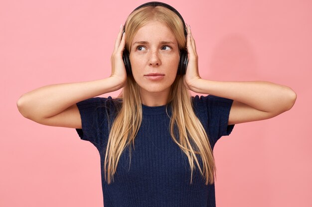 Isolated shot of cute teenage girl holding hands on ears, enjoying high quality music tracks in mp3 player using wireless headset