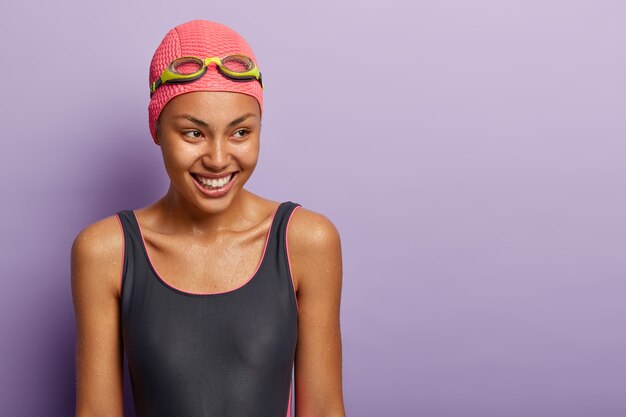 Free photo isolated shot of dark skinned woman athelete covers distance swimming, has healthy dark wet skin, wears swimsuit, goggles and hat, looks gladfully aside, trains in swimming pool. recreation time