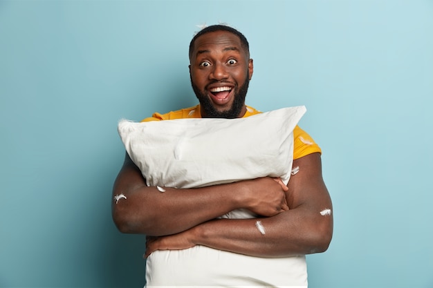 Isolated shot of dark skinned young man with optimistic gaze carries pillow, has fun in bedroom, enjoys comfort and peaceful domestic atmosphere at home, expresses good emotions. bed time concept