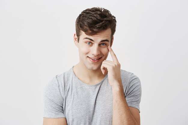Isolated shot of delighted cheerful young caucasian guy looking pleased, keeping finger on his temple, being glad to recieve big profit from sales, smiling with teeth, proud of himself.