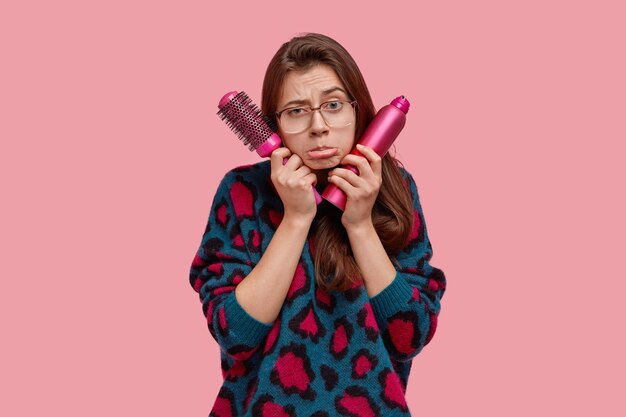 Isolated shot of depressed European woman carries hairbrush and spray, purses lower lip in discontent