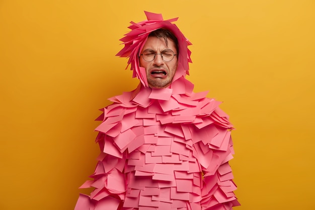 Isolated shot of displeased European man cries from despair, has difficult life and many problems, being sad and troubled, wears optical glasses and pink stickers, isolated on yellow wall