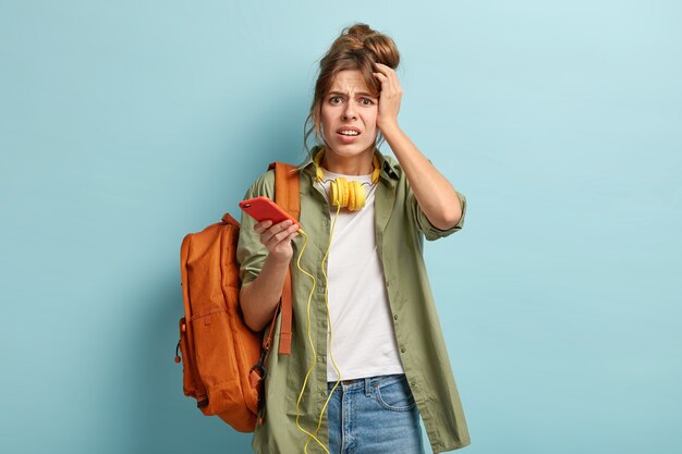 Isolated shot of dissatisfied schoolgirl holds cell phone connected to headphones