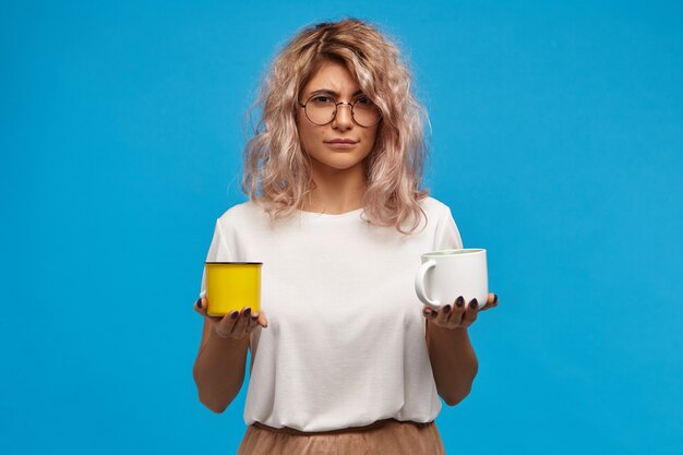 isolated shot of doubtful uncertain attractive young woman wearing stylish clothes holding white cup of hot chocolate or cocoa and yellow mug with warm milk, hesitating