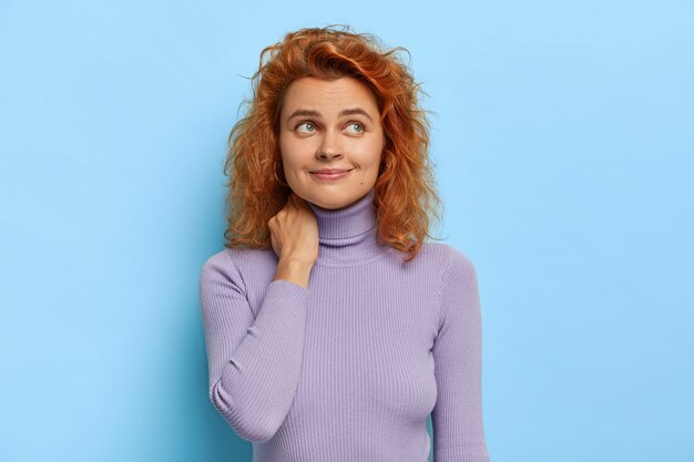 Isolated shot of dreamy curly haired ginger woman has satisfied thoughtful look, concentrated aside, wears purple polo neck jumper, recalls first date with boyfriend, isolated over blue wall