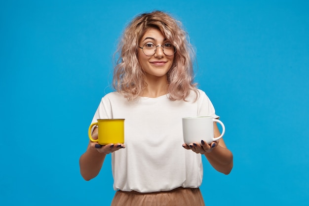 Isolated shot of fashionable charming young European woman in stylish round eyewear and oversize t-shirt having friendly look, holding two cups of hot drinks