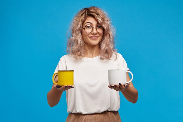 isolated shot of fashionable charming young european woman in stylish round eyewear and oversize t-shirt having friendly look, holding two cups of hot drinks