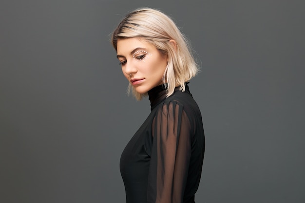 Isolated shot of fashionable trendy hipster with nose ring and blonde bob hairdo wearing transparent black blouse posing at blank wall, having thoughtful facial expression, looking down