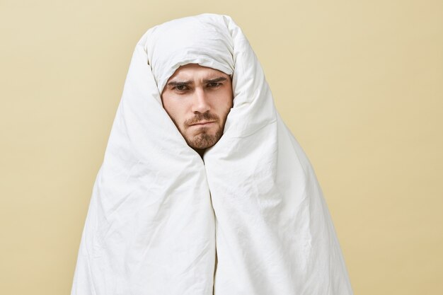 Isolated shot of frustrated sleepy young male having angry displeased facial expression frowning eyebrows, unwilling to got to work, preferring staying in warm bed instead, wrapped in white blanket