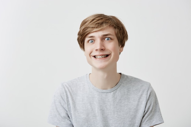 Foto gratuita il colpo isolato del giovane divertente serra i denti con le parentesi graffe, facendo i fronti, divertendosi con gli amici. ragazzo caucasico positivo con sorrisi di capelli chiari.