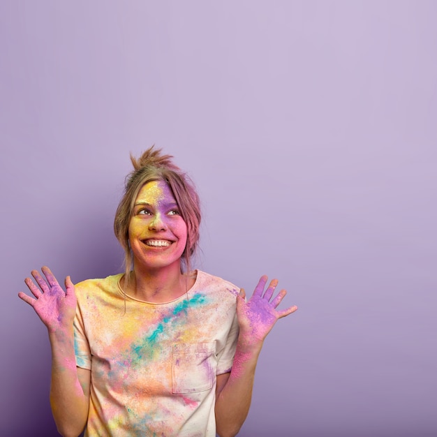 Free photo isolated shot of glad dreamy young woman focused upwards, raises both hands and shows colored palms, smiles positively, celebrates holi festival of colors, free space above for your information