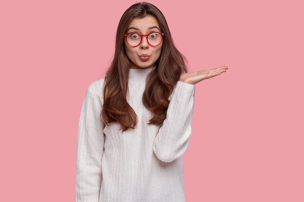 Isolated shot of good looking friendly experienced seller raises hand, demonstrates something against pink background, dressed in casual white sweater