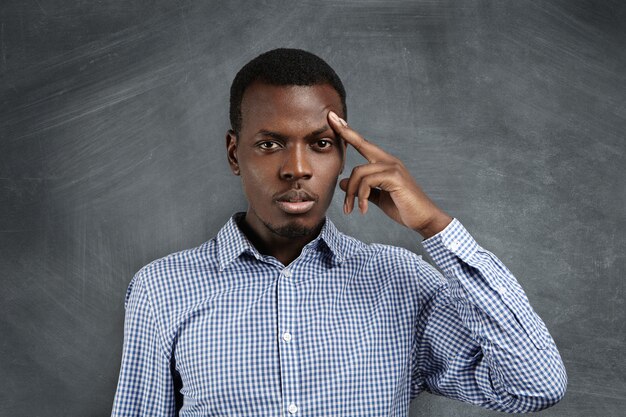 Isolated shot of handsome frustrated African man wearing shirt holding finger on his forehead as if trying hard to recollect something or solve serious problem, having concentrated look.