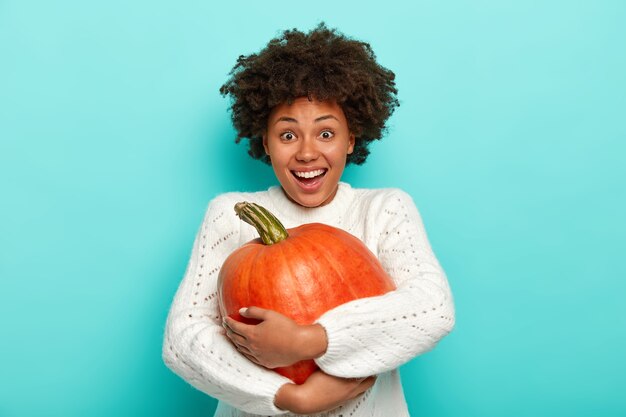 Isolated shot of happy Afro woman enjoys autumn season, holds big ripe pumpkin, picks up vegetable from autumnal garden, has joyful expression, wears white sweater, models against blue background