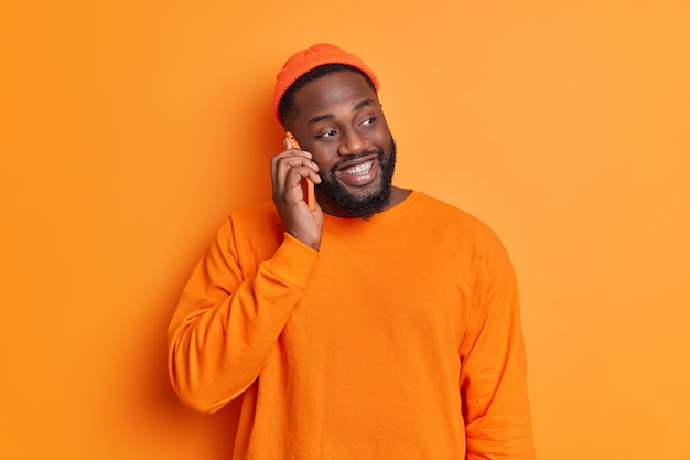 Isolated shot of happy bearded man has cheerful talk via mobile phone concentrated aside smiles joyfully wears hat and sweater poses against orange studio wall