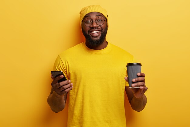 Isolated shot of happy black man in yellow clothing, types message, downloads new application on smart phone, enjoys coffee from disposable cup, has toothy smile, white teeth, thick bristle.