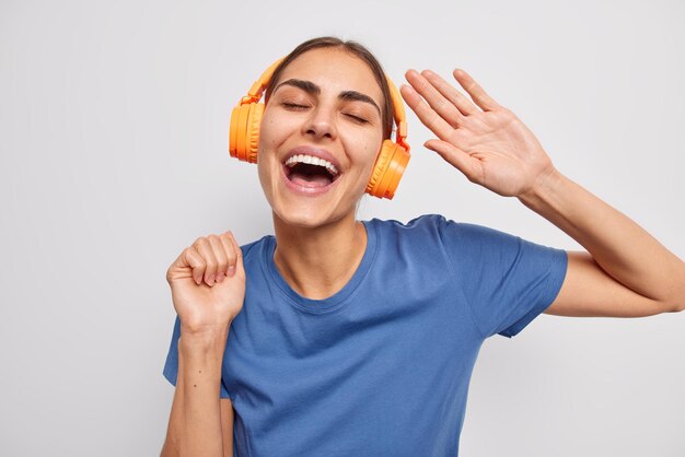 Isolated shot of happy dark haired woman smiles broadly enjoys favorite music in headphones dances carefree dressed in casual blue t shirt isolated over white background People and hobby concept