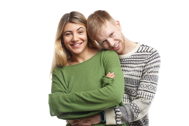 Isolated shot of happy handsome guy embracing gently his cute girlfriend. Stylish young engaged couple in love cuddling indoors, enjoying sweet moments of their happiness, smiling joyfully