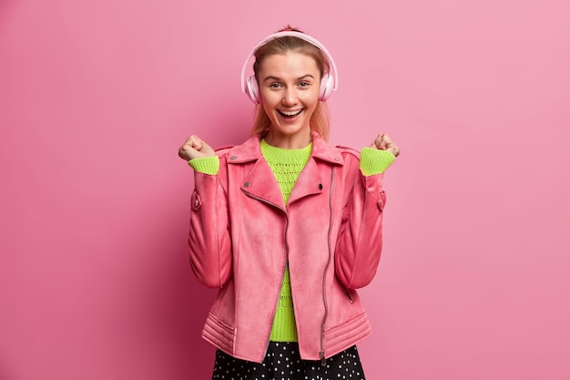 Foto gratuita il colpo isolato dell'adolescente felice ascolta la musica tramite le cuffie senza fili stereo solleva i pugni chiusi e sorride ampiamente