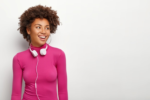 Colpo isolato di felice giovane donna afroamericana con capelli croccanti, distoglie lo sguardo con espressione felice, indossa orecchini e dolcevita rosa, isolato su sfondo bianco, utilizza cuffie stereo