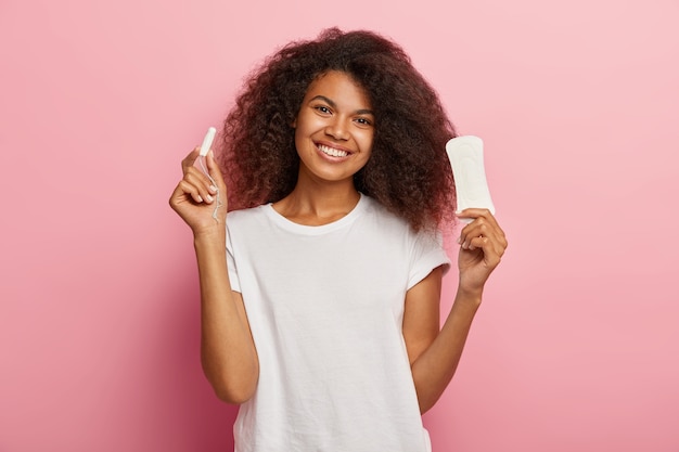 Isolated shot of happy young Afro female holds menstuation cotton tampon and sanitary napkin