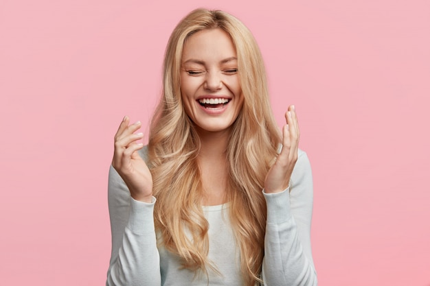 Isolated shot of joyful blonde young cute woman laughs joyfully as hears funny anecdote from friend, has long light hair, poses against pink  wall. Happiness and positive emotions concept