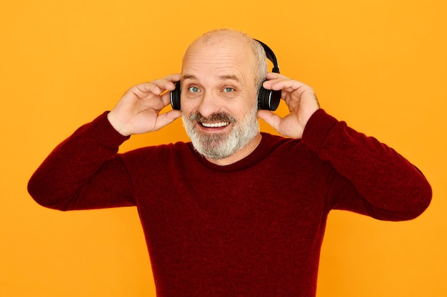 Colpo isolato di gioioso uomo anziano caucasico bello con la testa calva e la barba grigia che sorride assumendo wireless moderno collegandoli al gadget elettronico tramite bluetooth.