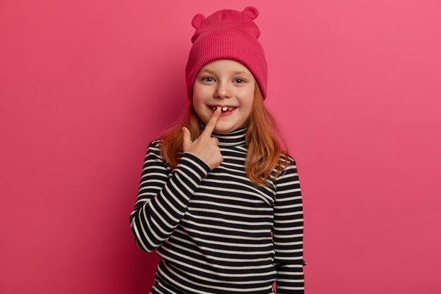 Isolated shot of little child rejoices having new adult tooth, has ginger hair, dresed in striped sweater and stylish hat, has charming positive expression poses over rosy wall. Look at my white teeth