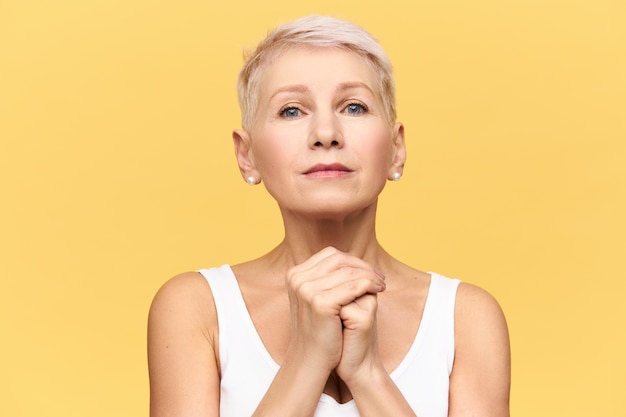 Isolated shot of modern blonde European mature woman in tank top keeping hands under her chin staring  with nervous upset look, eyes full of hope, begging, pleading, worrying about something