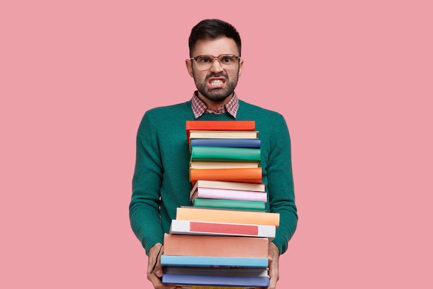 Изолированный снимок раздраженного молодого человека, стиснувшего зубы от гнева, держит огромную стопку книг, носит очки и зеленый свитер, делает домашнее задание