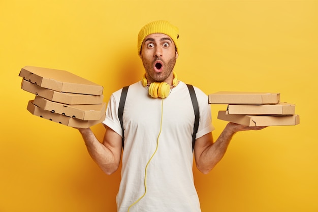 Изолированный снимок удивленного доставщика, держащего в руках несколько картонных коробок с итальянской пиццей, шокированного доставкой фаст-фуда в неправильное место, белой футболки и наушников на шее