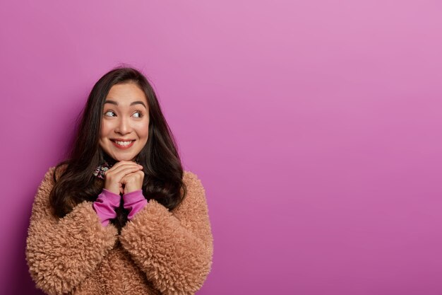 Isolated shot of optimistic Japanese woman with hands pressed under chin, wears brown fur coat, thinks about pleasant moment in life