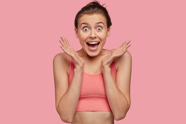 Isolated shot of overjoyed smiling young female fitness model in pink top clasps hands