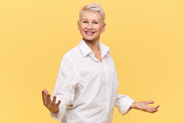Isolated shot of overjoyed successful mature woman boss in white shirt holding hands wide apart and smiling happily, giving motivational speech, energizing employees, her posture expressing confidence