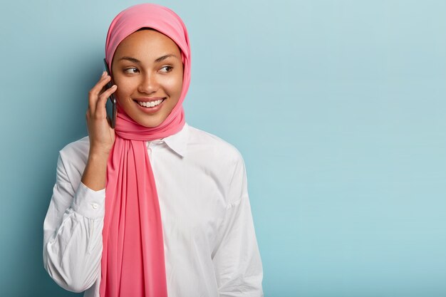 유쾌한 여성 모델의 고립 된 샷은 현대 휴대 전화를 통해 말하고, 누군가와 이야기하고, 진심으로 미소를 짓고, 멀리 집중하고, 빈 공간이있는 파란색 벽에 서서, 이슬람 옷을 입습니다.