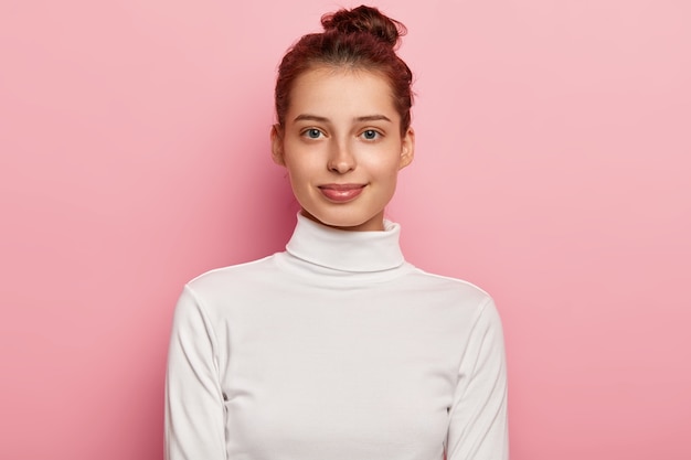 Isolated shot of pleasant looking young woman wears white turtleneck, has hair bun, looks with calm facial expressions, poses against pink background.