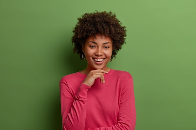 Isolated shot of pleased dark skinned Afro American woman keeps hand under chin, smiles gently, wears casual rosy sweater, poses against green wall, hears something funny, looks pleased