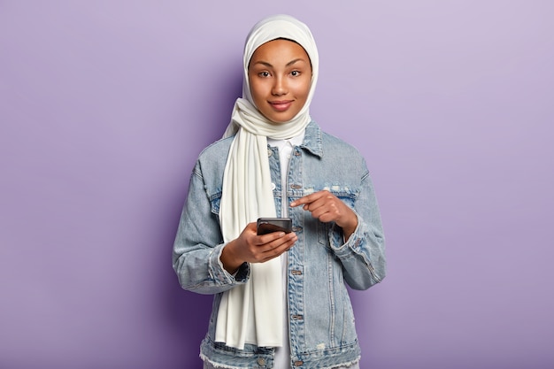 Free photo isolated shot of pleased mixed race young woman with dark skin, follows muslim religion, points at mobile phone screen, asks to read internet news on website, isolated over purple wall.