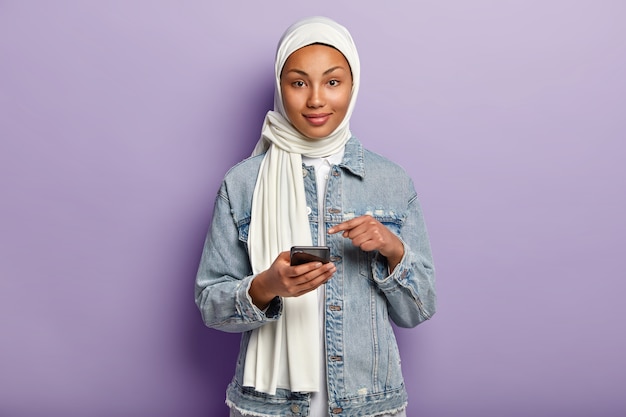 Изолированный снимок довольной молодой женщины смешанной расы с темной кожей, исповедующей мусульманскую религию, указывает на экран мобильного телефона, просит читать интернет-новости на веб-сайте, изолированном над фиолетовой стеной.