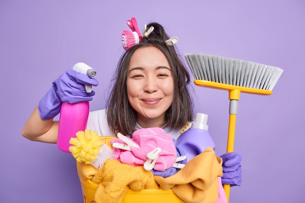 Isolated shot of positive brunette asian housewife holds detergent for cleaning holds broom poses near laundry basket does disinfection of house isolated over purplebackground. cheerful housekeeper