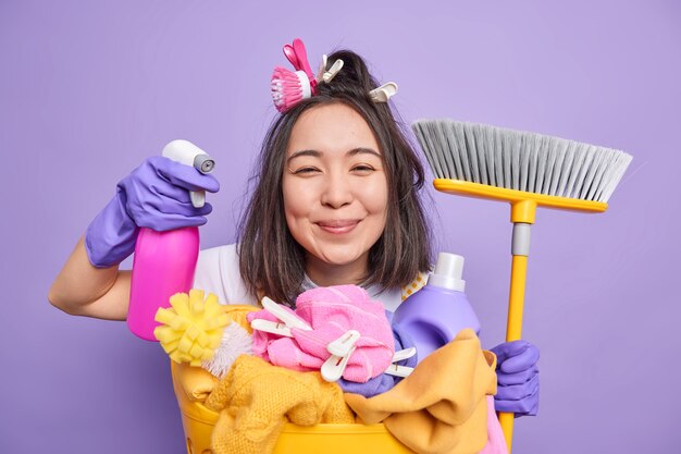 ポジティブブルネットのアジアの主婦の隔離されたショットは、洗濯かごの近くでほうきのポーズを保持するクリーニングのための洗剤を保持し、紫の背景の上に隔離された家の消毒を行います。陽気な家政婦