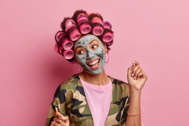 Isolated shot of positive woman looks aside smiles broadly raises arm applies hair rollers beauty mask dressed in domestic robe prepares for special occasion wants to look beautiful models indoor