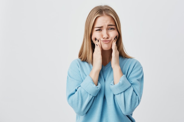 Isolated shot of pretty cute blonde girl in blue clothes touching her cheeks with her hands, showing how big they are. Funny female mocking and posing.