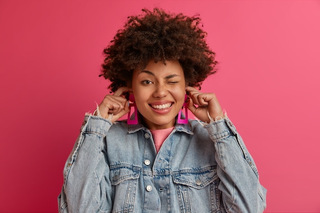 Free photo isolated shot of pretty dark skinned female blocks ears, avoids noise, winks eye, smiles broadly, asks to turn off volume, doesnt want to hear loud music, dressed in denim jacket, trendy earrings