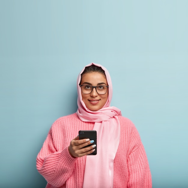 꽤 꿈꾸는 이슬람 여성의 고립 된 샷은 소셜 네트워크에서 메시지를 보내기 위해 휴대 전화를 사용하고 위쪽으로 집중하고 메시지 내용에 대해 생각하고 파란색 벽 위에 서 있습니다.